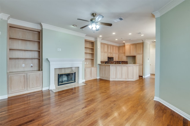 unfurnished living room featuring built in features, a tiled fireplace, ceiling fan, crown molding, and light hardwood / wood-style flooring