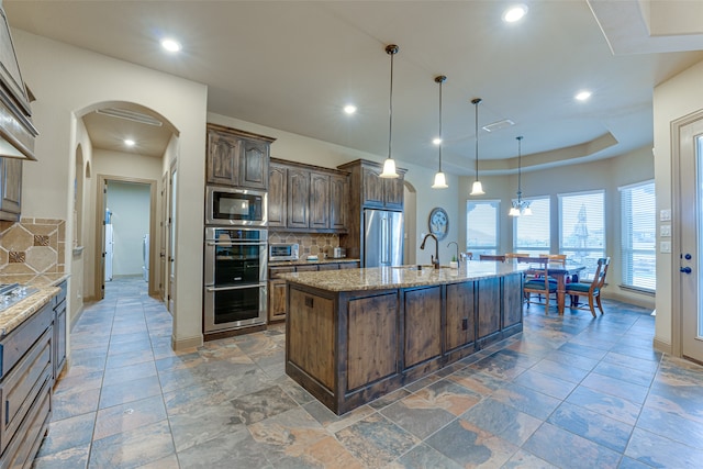 kitchen featuring light stone counters, tasteful backsplash, an island with sink, appliances with stainless steel finishes, and decorative light fixtures