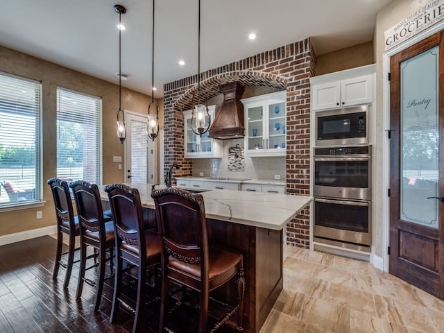 kitchen with stainless steel appliances, hanging light fixtures, an island with sink, custom range hood, and white cabinetry