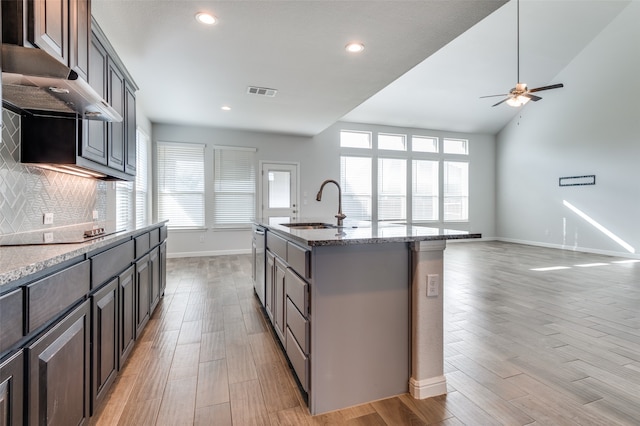 kitchen with decorative backsplash, light stone countertops, black electric cooktop, sink, and an island with sink