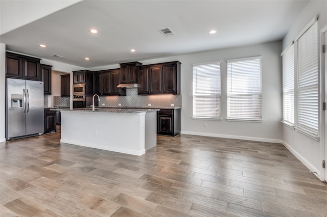 kitchen featuring light wood-type flooring, tasteful backsplash, dark brown cabinets, stainless steel appliances, and an island with sink