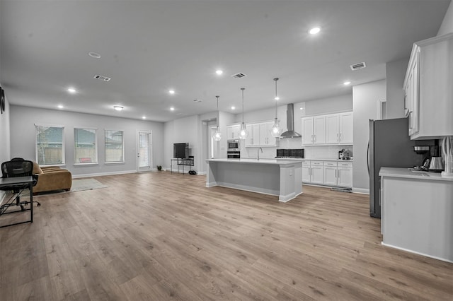 kitchen with light hardwood / wood-style floors, white cabinetry, decorative light fixtures, a kitchen island with sink, and wall chimney range hood