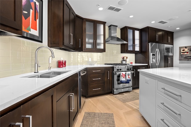 kitchen with wall chimney range hood, appliances with stainless steel finishes, backsplash, light wood-type flooring, and sink