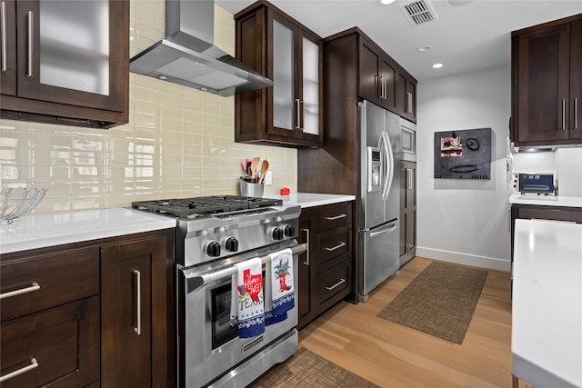 kitchen featuring tasteful backsplash, wall chimney range hood, dark brown cabinets, light wood-type flooring, and appliances with stainless steel finishes