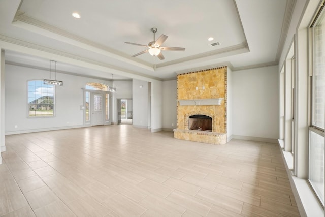 unfurnished living room with ceiling fan, a stone fireplace, crown molding, a tray ceiling, and light wood-type flooring