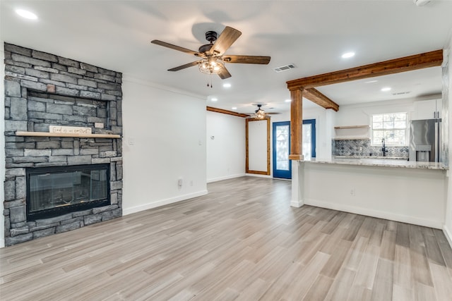 unfurnished living room featuring beamed ceiling, ornamental molding, ceiling fan, a stone fireplace, and light wood-type flooring