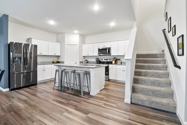 kitchen with stainless steel appliances, hardwood / wood-style floors, stone counters, an island with sink, and white cabinetry