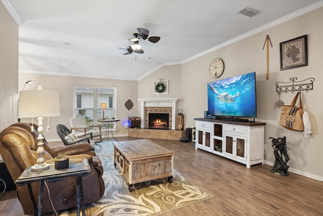 living room with ceiling fan, wood-type flooring, crown molding, a tile fireplace, and vaulted ceiling