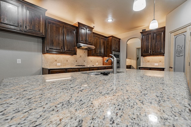 kitchen with decorative backsplash, hanging light fixtures, sink, light stone countertops, and dark brown cabinets