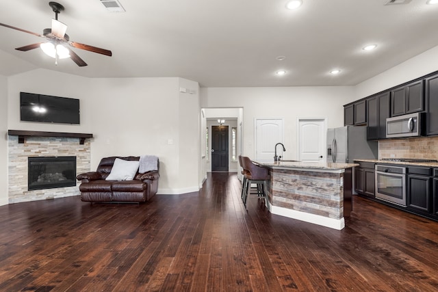 kitchen with dark wood-type flooring, light stone countertops, appliances with stainless steel finishes, and a center island with sink