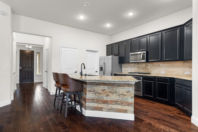 kitchen featuring dark wood-type flooring, an island with sink, backsplash, light stone countertops, and appliances with stainless steel finishes