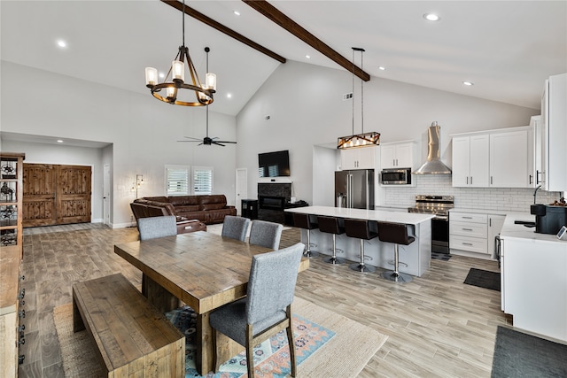 dining space with high vaulted ceiling, ceiling fan with notable chandelier, sink, light wood-type flooring, and beamed ceiling