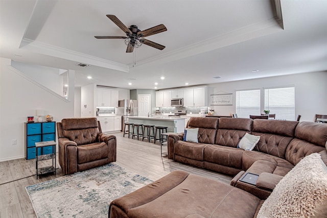 living room featuring a raised ceiling, ceiling fan, crown molding, and light wood-type flooring