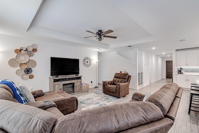 living room featuring light wood-type flooring, ornamental molding, a raised ceiling, ceiling fan, and a fireplace