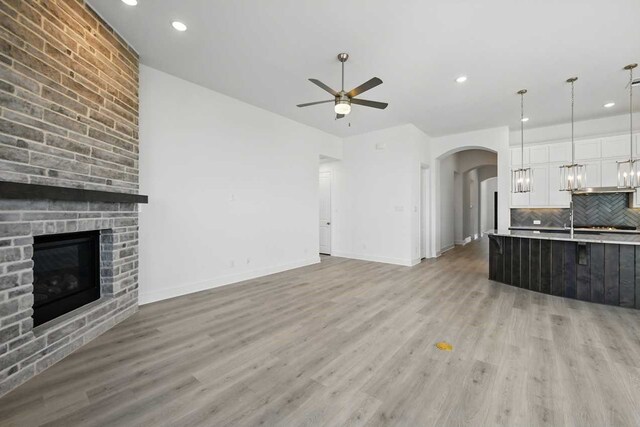 unfurnished living room with a large fireplace, baseboards, ceiling fan, arched walkways, and light wood finished floors