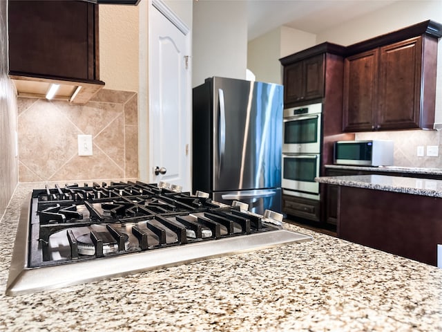 kitchen featuring dark brown cabinetry, decorative backsplash, light stone counters, and stainless steel appliances