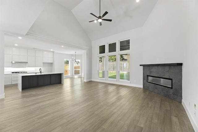 unfurnished living room with high vaulted ceiling, ceiling fan with notable chandelier, a fireplace, a sink, and light wood-type flooring