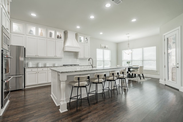 kitchen with stainless steel refrigerator, white cabinetry, pendant lighting, a center island with sink, and custom range hood