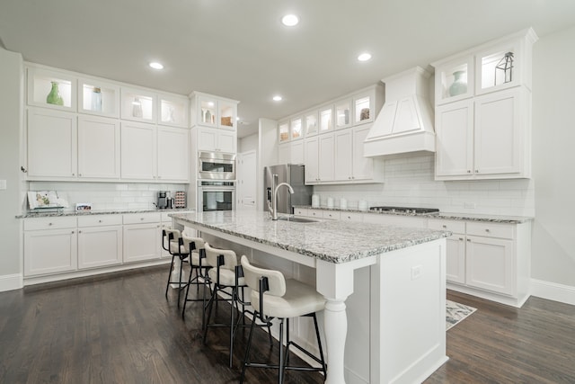 kitchen featuring white cabinets, stainless steel appliances, premium range hood, and a kitchen island with sink