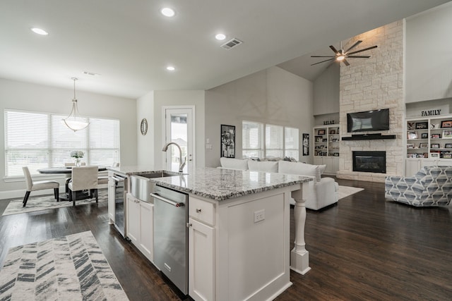 kitchen featuring stainless steel dishwasher, a center island with sink, white cabinets, a stone fireplace, and hanging light fixtures