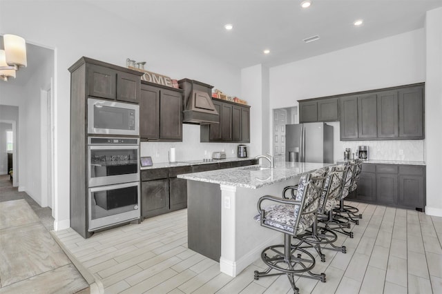 kitchen with custom exhaust hood, stainless steel appliances, light stone counters, a center island with sink, and a breakfast bar area