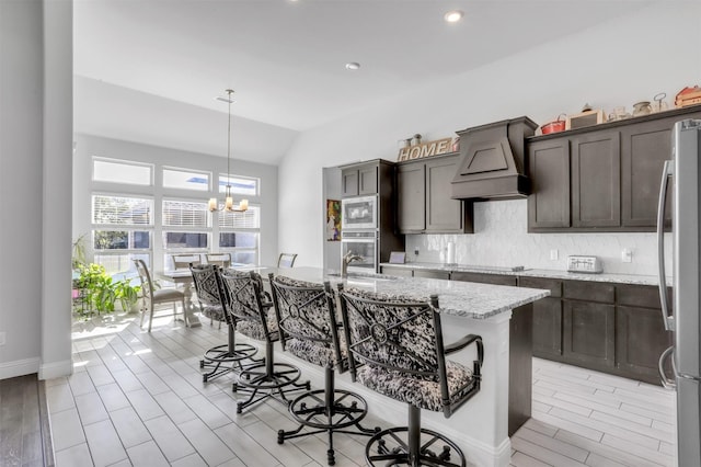 kitchen with appliances with stainless steel finishes, an island with sink, backsplash, light stone counters, and custom range hood