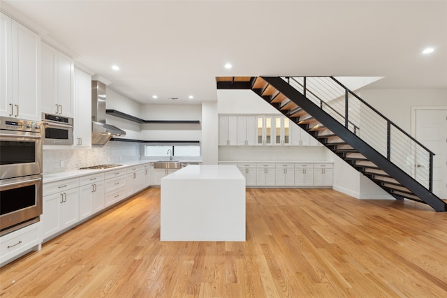 kitchen featuring a center island, white cabinets, wall chimney exhaust hood, double oven, and light wood-type flooring