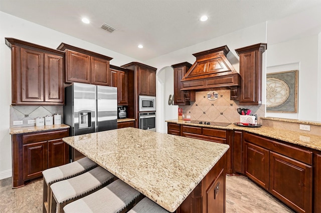 kitchen with a kitchen breakfast bar, a center island, light stone counters, stainless steel appliances, and custom range hood