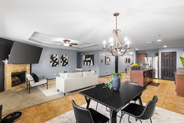 dining space featuring a tiled fireplace, ceiling fan with notable chandelier, and a tray ceiling