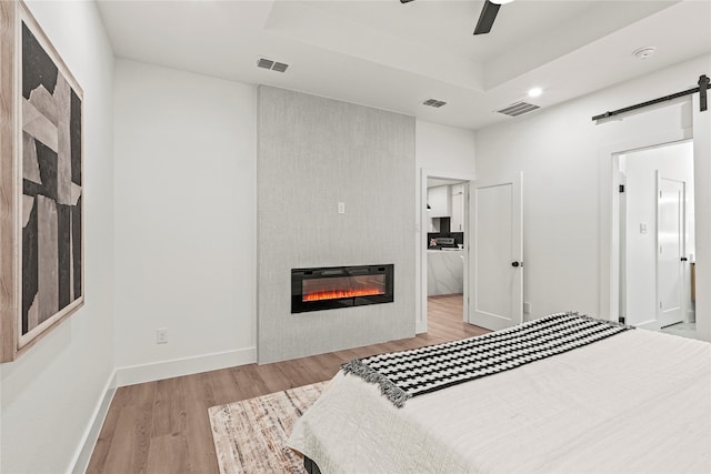 bedroom featuring a fireplace, a barn door, a raised ceiling, ceiling fan, and light hardwood / wood-style flooring