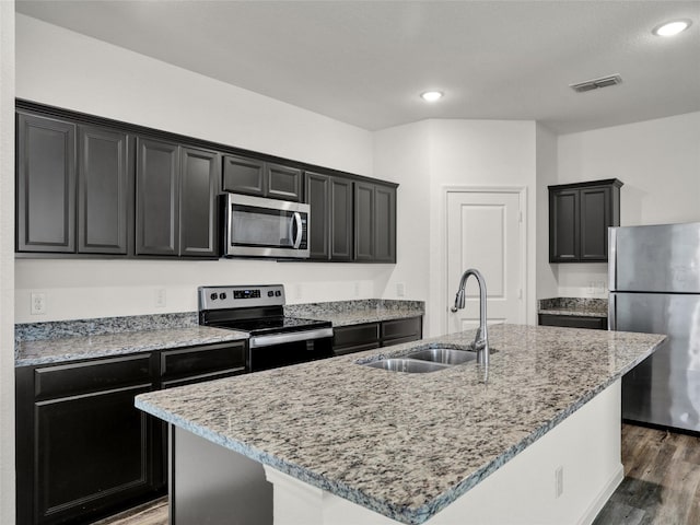 kitchen with sink, a kitchen island with sink, stainless steel appliances, light stone countertops, and dark hardwood / wood-style flooring