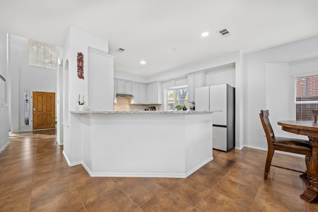 kitchen featuring light stone counters, white refrigerator, kitchen peninsula, tasteful backsplash, and white cabinetry