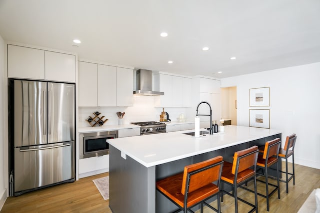 kitchen with appliances with stainless steel finishes, a kitchen bar, an island with sink, wall chimney exhaust hood, and light hardwood / wood-style flooring