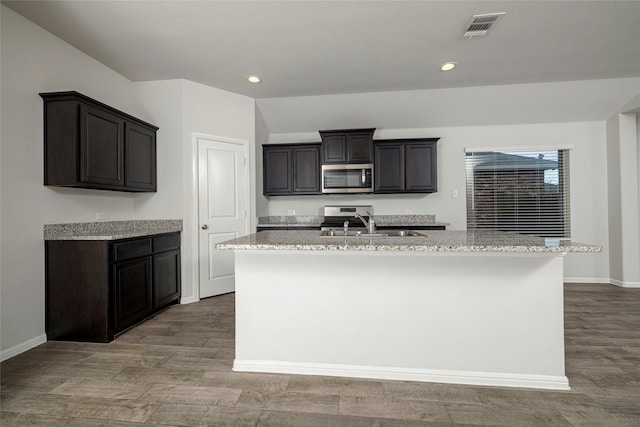 kitchen with hardwood / wood-style flooring, light stone counters, an island with sink, and stainless steel appliances