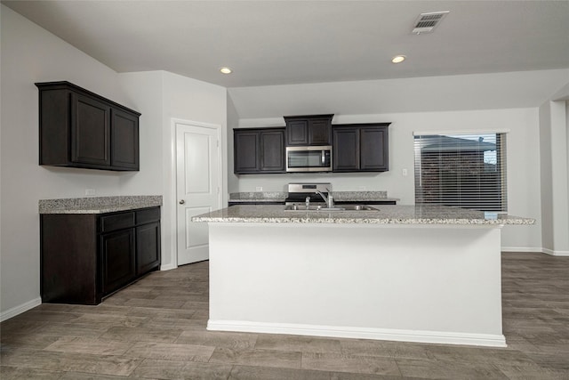 kitchen with an island with sink, hardwood / wood-style flooring, light stone countertops, sink, and appliances with stainless steel finishes