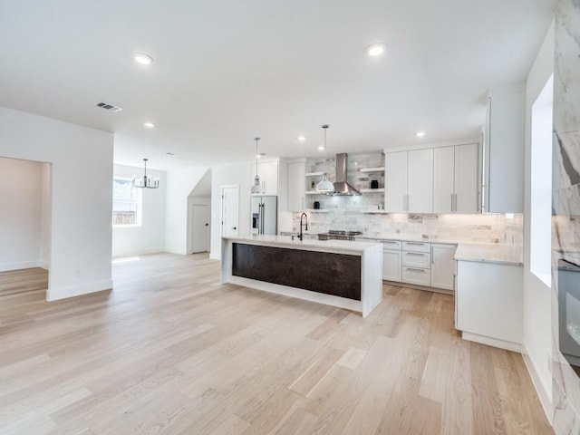 kitchen with a kitchen island with sink, white cabinets, stainless steel refrigerator with ice dispenser, hanging light fixtures, and wall chimney exhaust hood