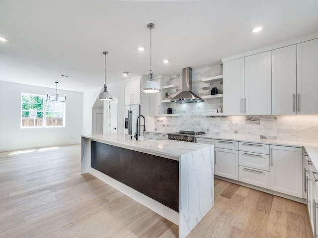 kitchen with light stone counters, wall chimney exhaust hood, decorative light fixtures, a center island with sink, and white cabinetry