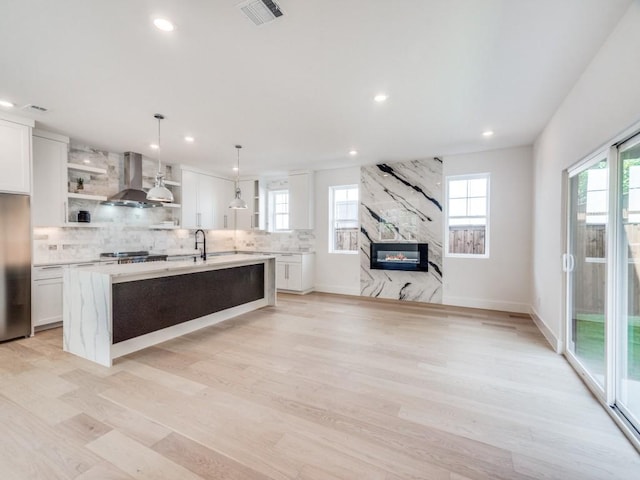 kitchen with white cabinets, a center island with sink, hanging light fixtures, wall chimney exhaust hood, and light wood-type flooring