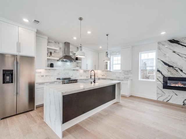 kitchen featuring white cabinets, hanging light fixtures, wall chimney exhaust hood, appliances with stainless steel finishes, and light stone counters