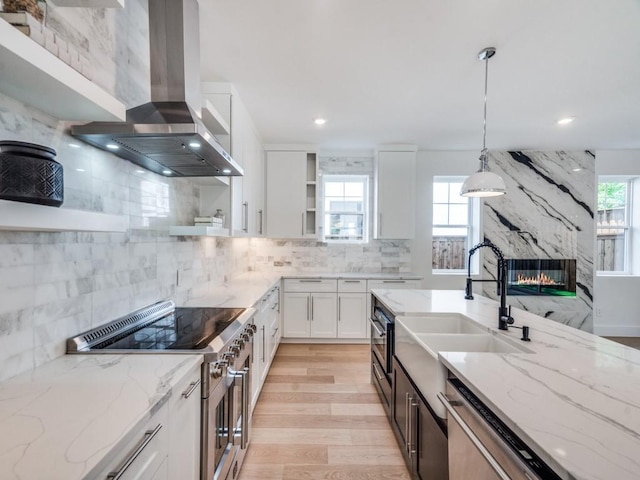 kitchen with wall chimney exhaust hood, light stone counters, decorative light fixtures, white cabinets, and appliances with stainless steel finishes