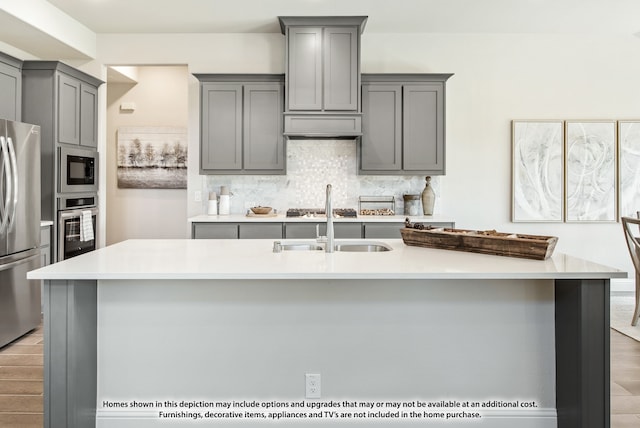 kitchen with decorative backsplash, gray cabinetry, a kitchen island with sink, light wood-type flooring, and appliances with stainless steel finishes