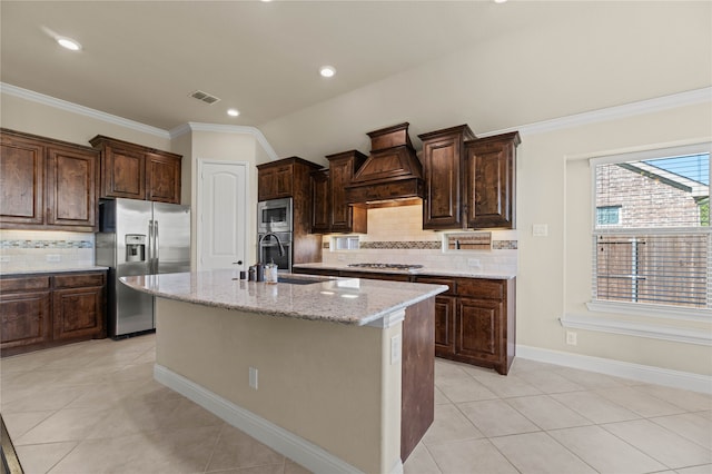 kitchen with tasteful backsplash, premium range hood, stainless steel appliances, sink, and a center island with sink