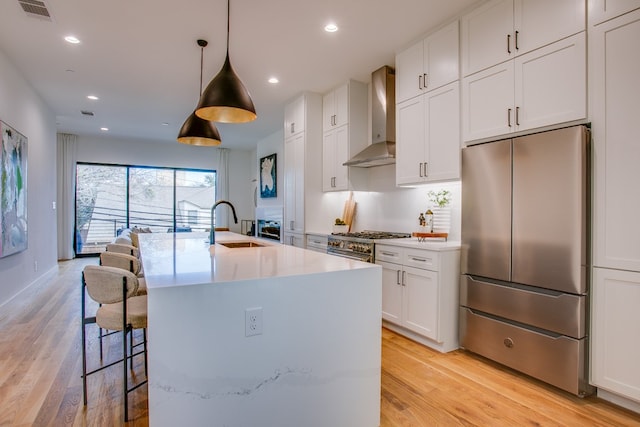 kitchen with light hardwood / wood-style floors, high end appliances, an island with sink, wall chimney range hood, and pendant lighting