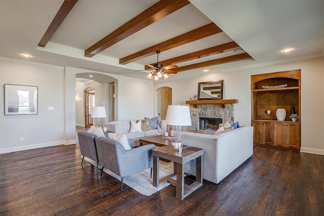 living room featuring ornamental molding, ceiling fan, dark hardwood / wood-style floors, beam ceiling, and a stone fireplace