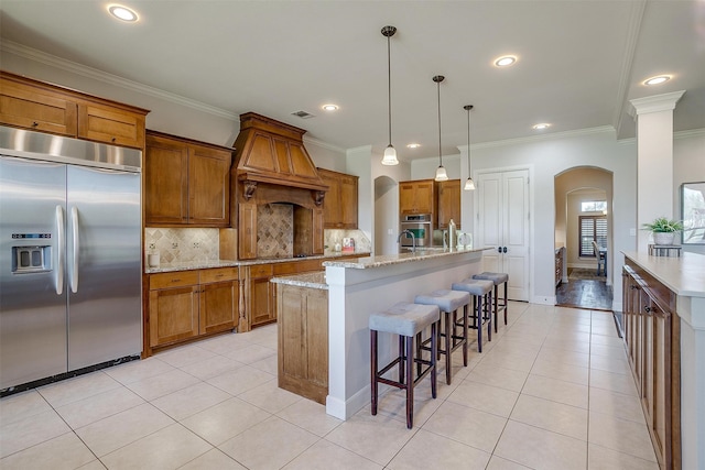 kitchen featuring appliances with stainless steel finishes, custom range hood, decorative light fixtures, a kitchen island with sink, and crown molding