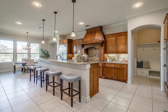 kitchen with tasteful backsplash, built in fridge, pendant lighting, custom exhaust hood, and a kitchen island with sink