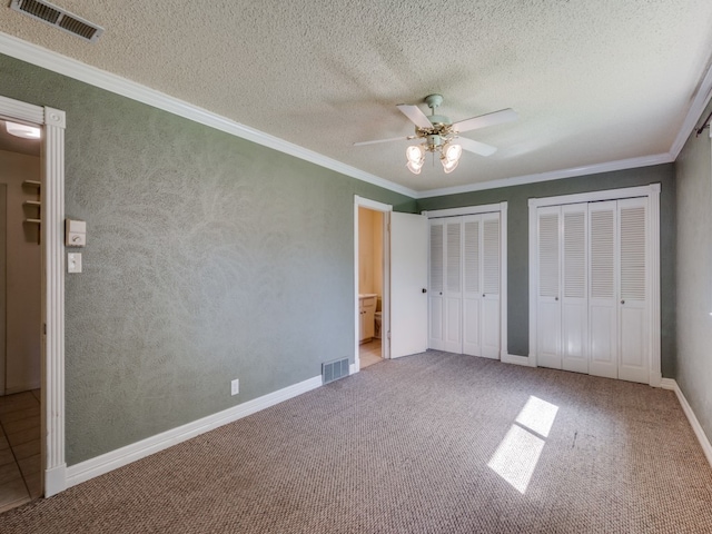 unfurnished bedroom with carpet flooring, a textured ceiling, ornamental molding, ceiling fan, and multiple closets