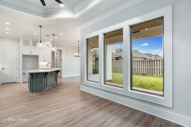 kitchen with crown molding, light wood-type flooring, hanging light fixtures, white cabinets, and a kitchen island with sink