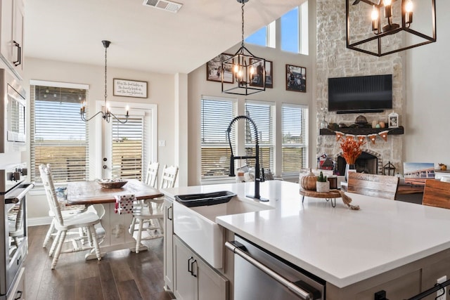 kitchen with a stone fireplace, a center island with sink, dark hardwood / wood-style floors, appliances with stainless steel finishes, and decorative light fixtures