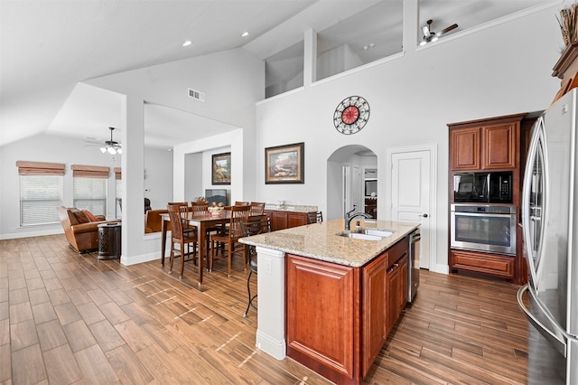 kitchen with wood-type flooring, light stone counters, a center island with sink, stainless steel appliances, and high vaulted ceiling